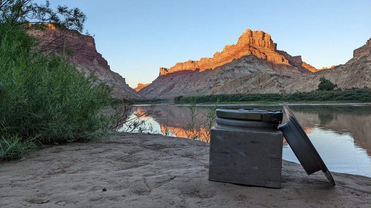 a self-contained toilet or "groover" on a river bank with a view of the river and red rock formations at sunset