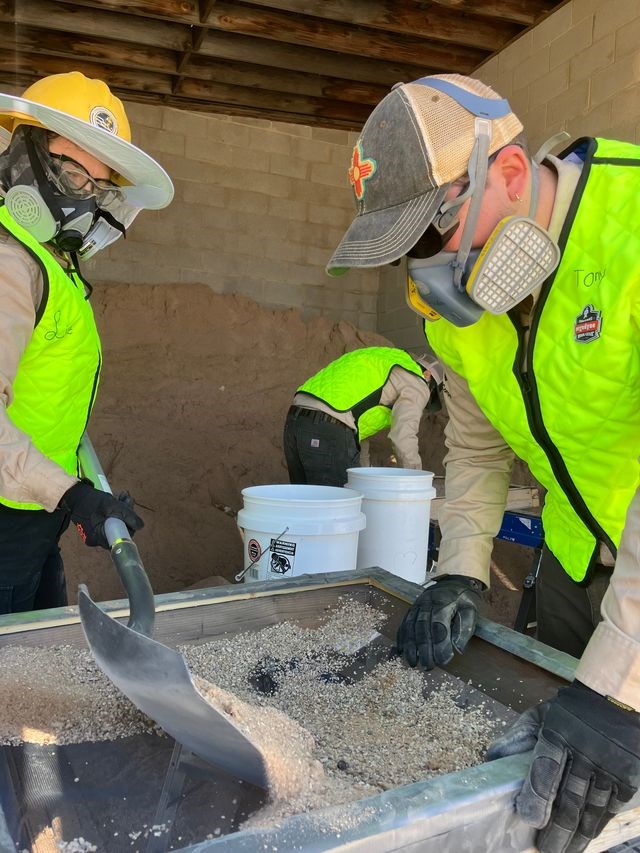 Three people wearing green reflective vests, tan long sleeve shirts, black work gloves, safety glasses and respirators. The two in the front are shoveling coarse sand onto a mesh screen. The third is setting up another screen in the background. They are i