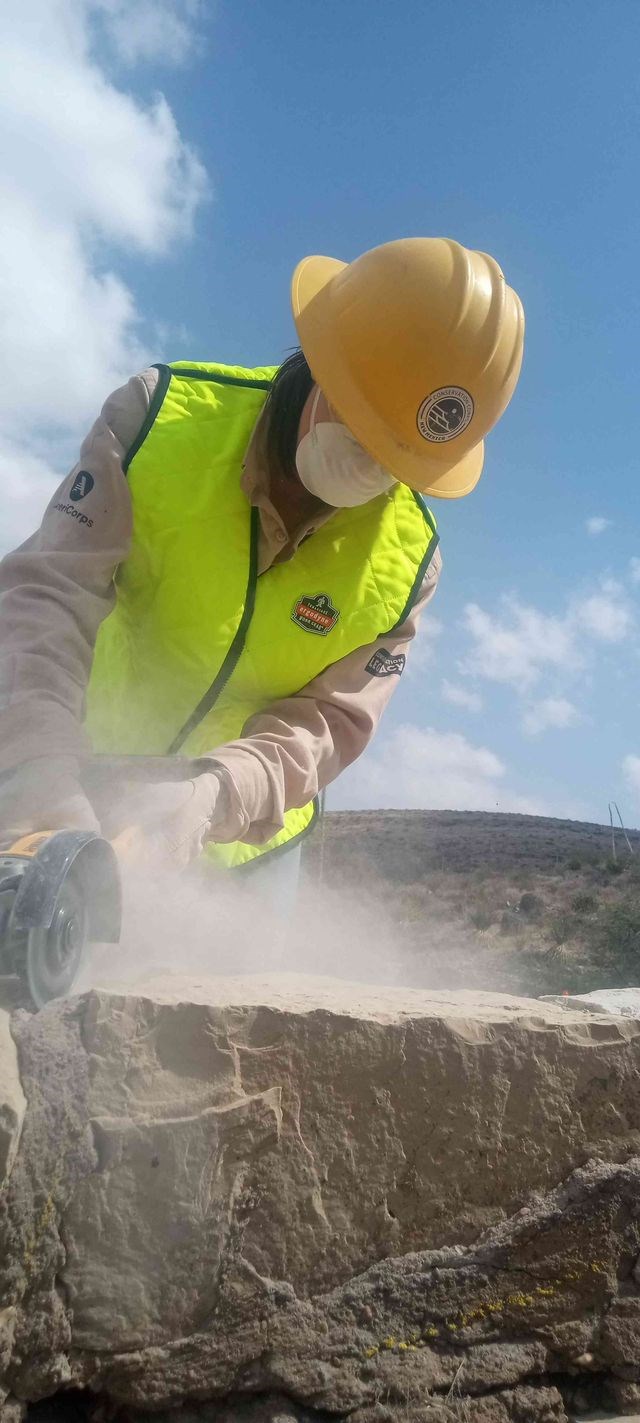 A person is outside wearing a tan long sleeve shirt, white dust mask and a yellow safety vest. The historic stone guardrail is in the foreground. They are using a small handheld grinder to remove old cement mortar from between two rocks.