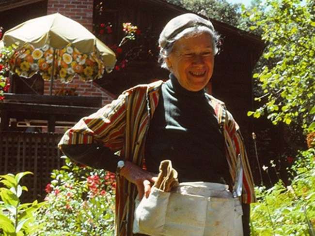 Smiling woman with gardening belt stands in lush garden on sunny day, wooden home with brick chimney behind her.