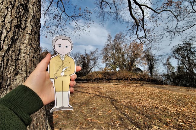 A cartoon illustration cutout of Alexander Hamilton in front of Redoubt 10, a mound fortified by wooden spikes.