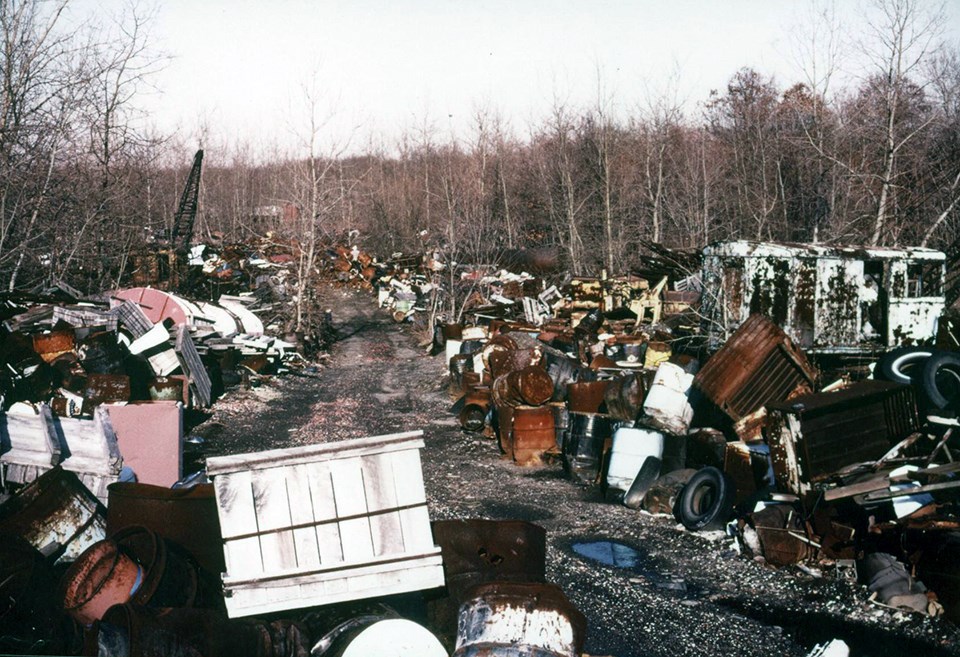 Metal waste, tires, and other garbage in giant piles in a clearing in a forest.