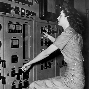 A young woman with dark curly hair wearing work pants and a work shirt. The woman is sitting on a stool staring at a wall full of meters and dials.