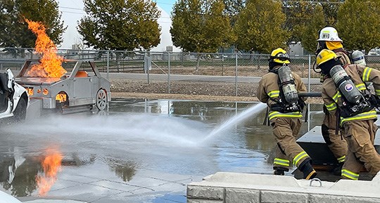 Structural firefighters carry a hose toward the source of a fire from a car fire training apparatus.