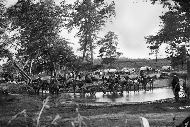 horse-drawn artillery crossing a shallow stream in front of a gathering of soldiers and a collection of covered wagons.