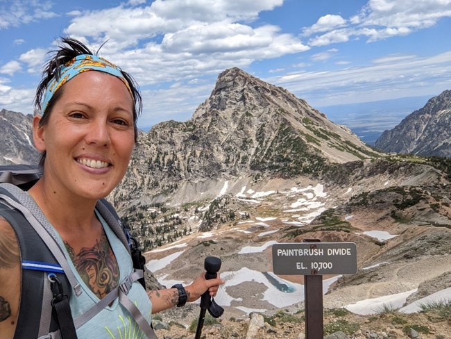 Woman on a hike smiles while standing near a sign reading Paintbrush Divide, with rugged mountain scenery behind her.