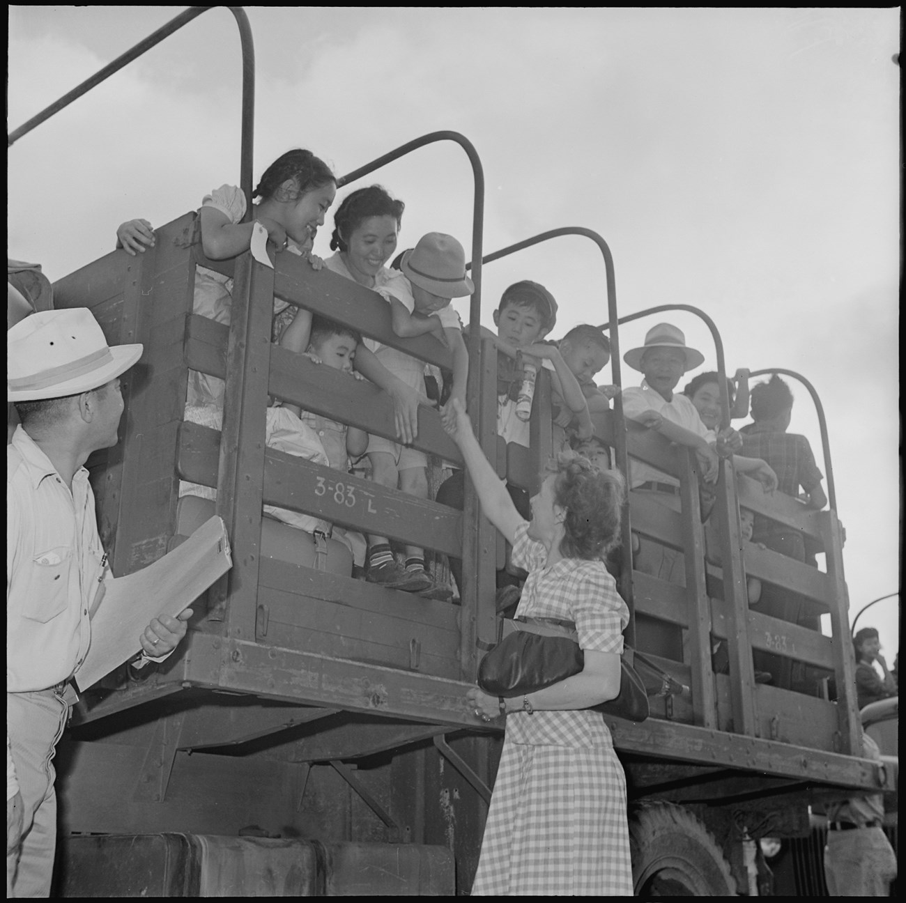 White teacher reaches up toward Japanese American children on the back of a crowded truck bed. The children smile down at her and reach over the ledge to say goodbye.
