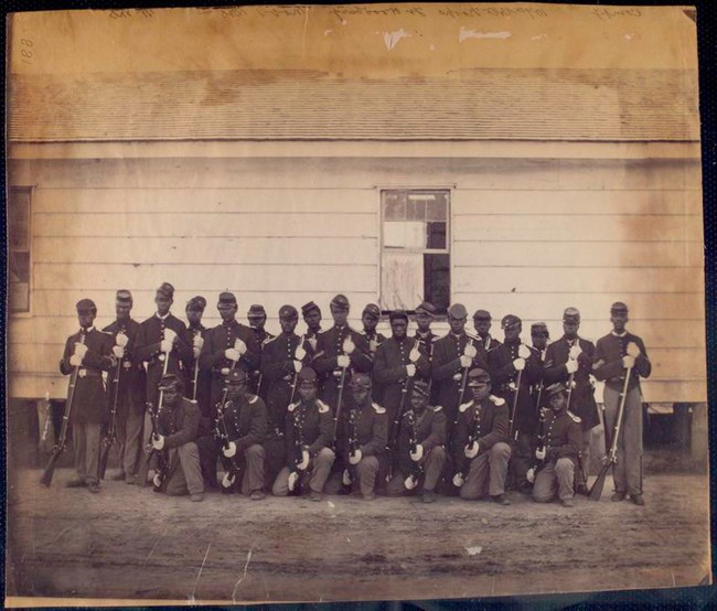 Line of black soldiers standing uniform while holding weapons at attention in front of a white house