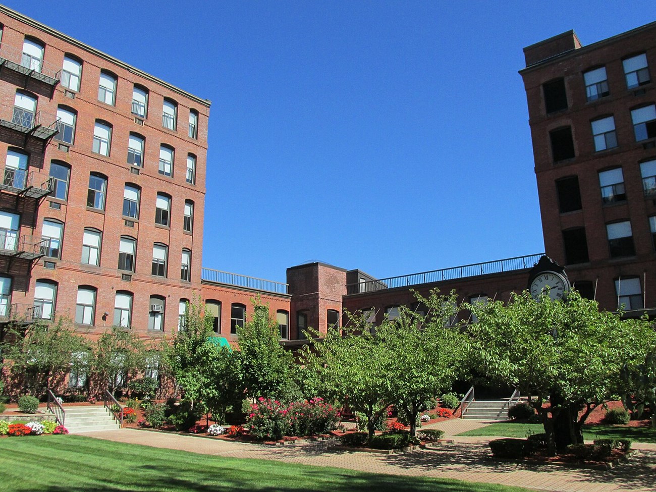 Six story red brick building on the left and right connected by a one story brick building, almost entirely covered by green plants and flowers in a courtyard.
