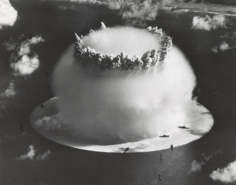 From above, ocean explosion surrounded by enormous ball of condensation with crown of smoke, a dozen dark battleships appearing like rice grains in comparison.