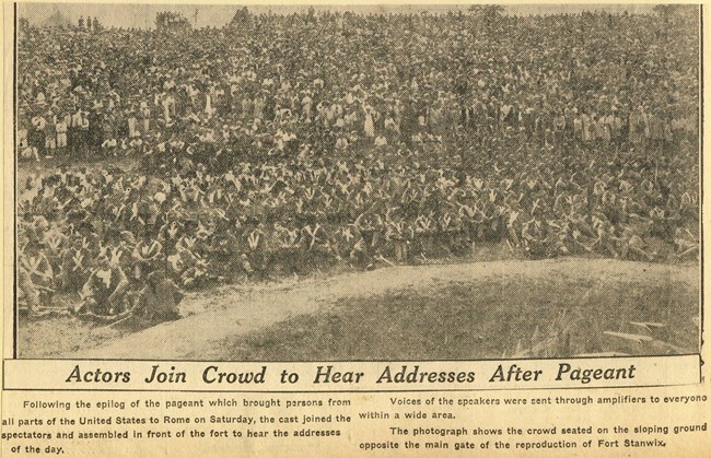 Faded newsprint picture of hundreds of people in a crowd, peering at a miniature fort while a performance is enacted around it.