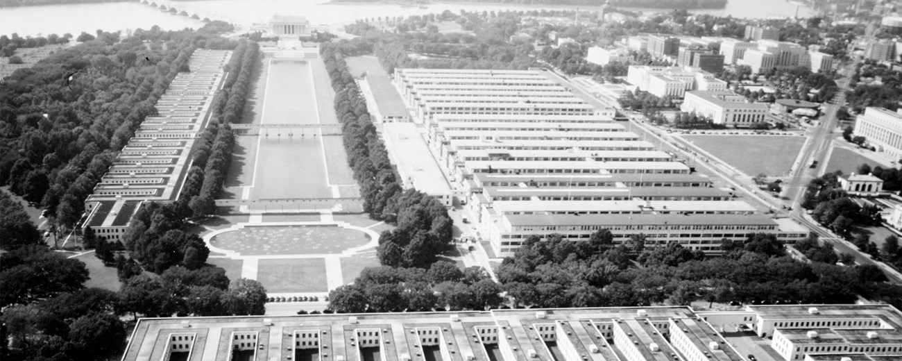 Black and white aerial photo of a city showing several buildings, a large greenspace, and a river in the background.