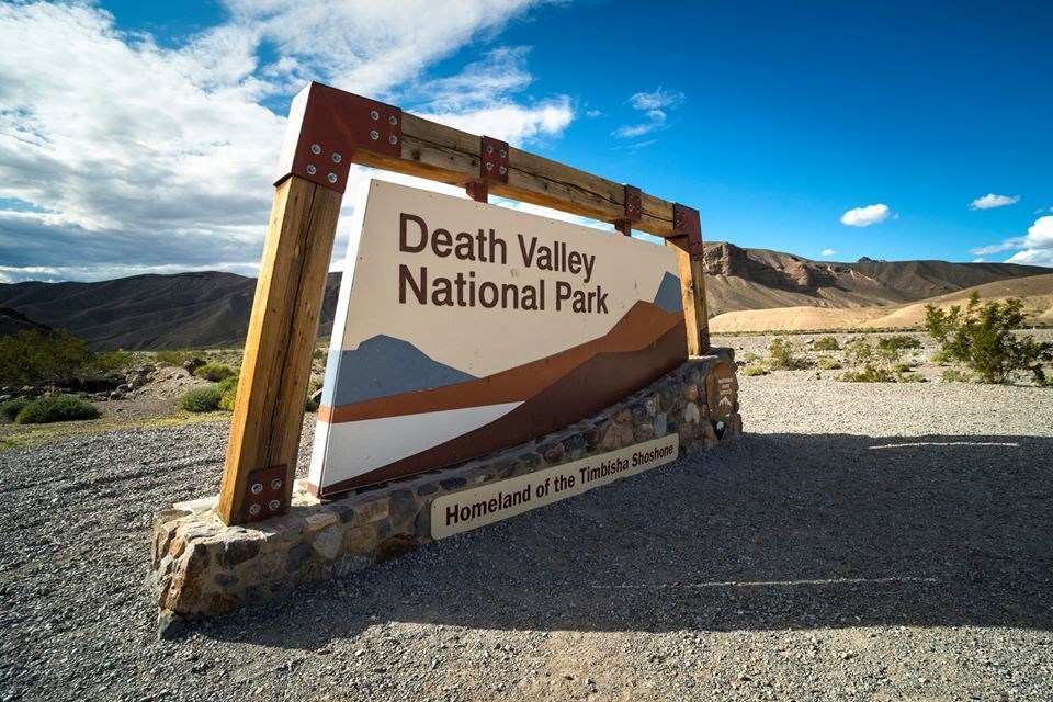 A large sign that reads "Death Valley National Park: Homeland of the Timbisha Shoshone" with a wooden border and stone foundation, with brown hills and a partly cloudy blue sky in the background.