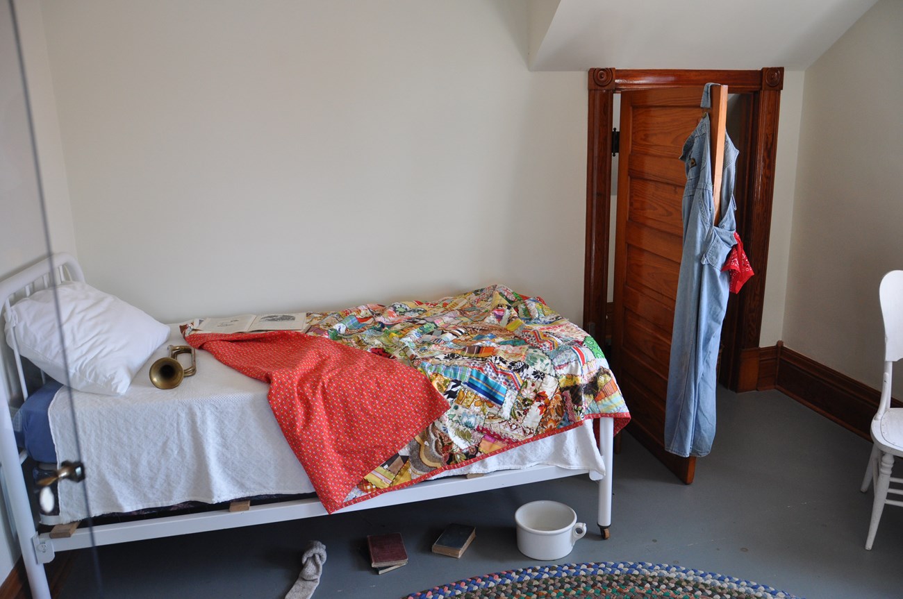 A twin bed with a white frame and colorful blanket with dirty socks on the floor, trumpet on the bed, and overalls hanging on the closet door.