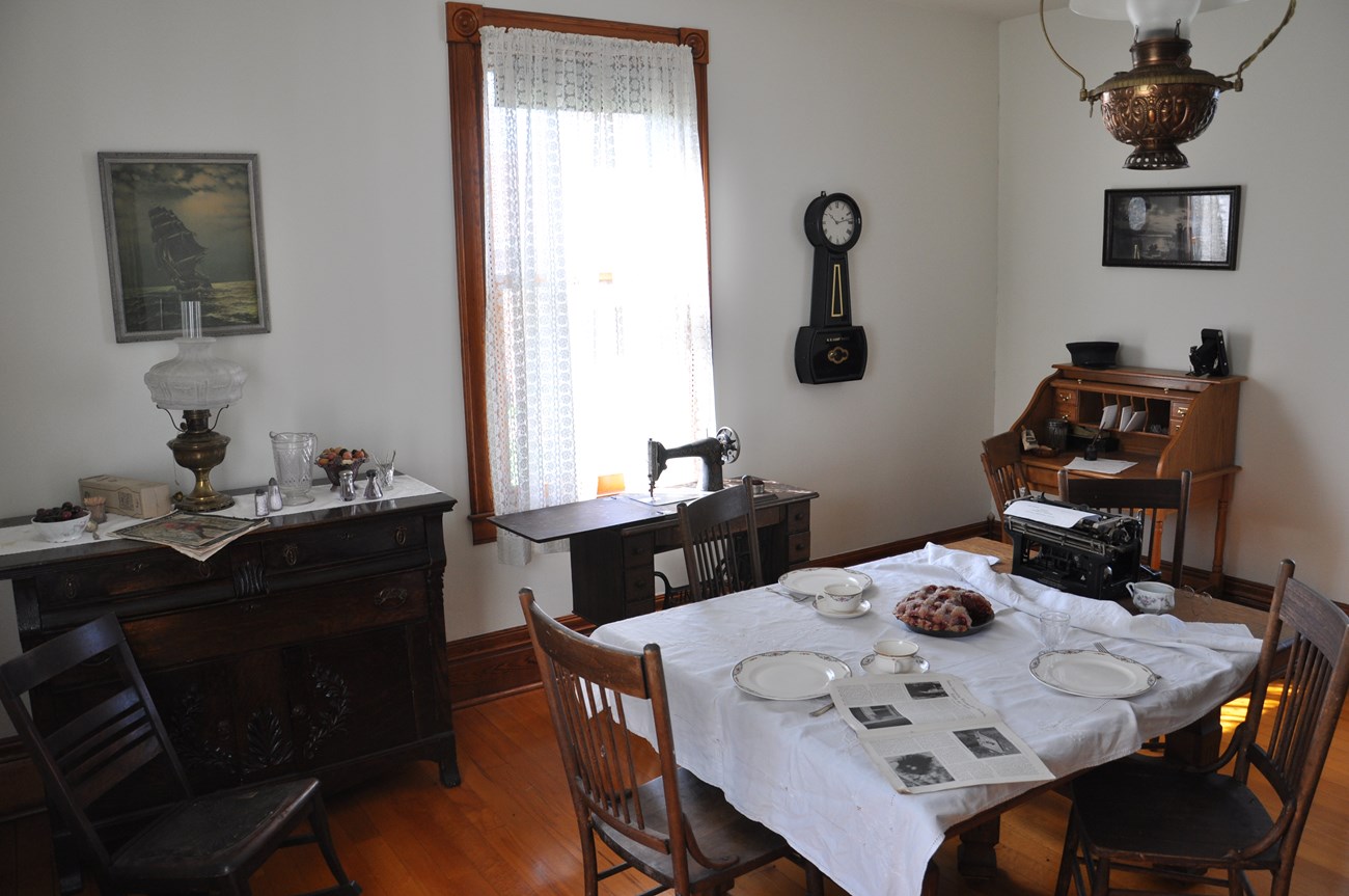 A dining room table set with plates, cups, a pie, and type writer. An antique Singer sewing machine in the back ground and clock on the wall.
