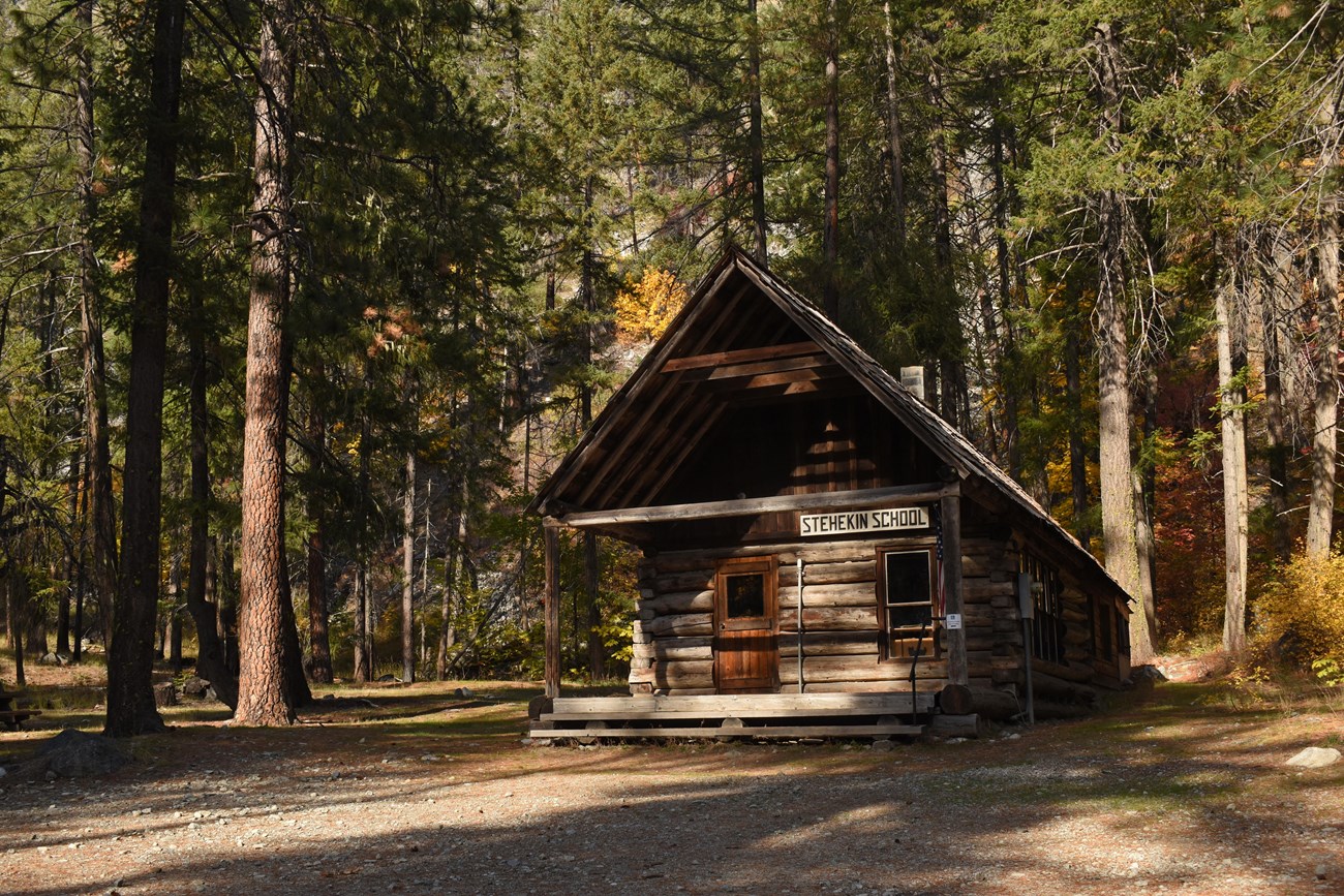 The Stehekin School A wooden one-room school building with an American flag on a pole on the porch, sits in a sparse forest.