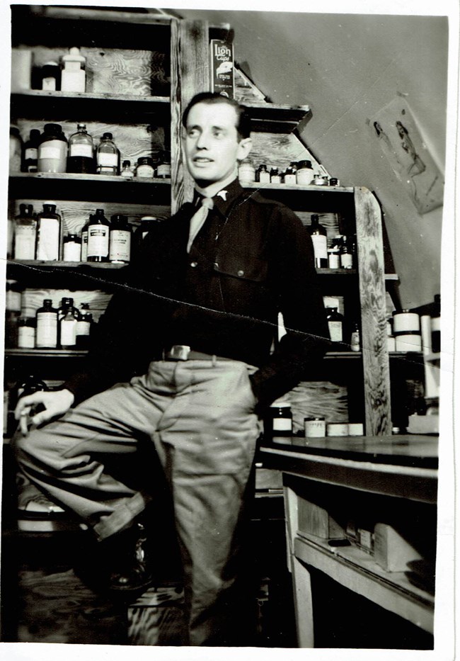 Black and white photo of a man sitting on a stool in a small room filled with medicine bottles
