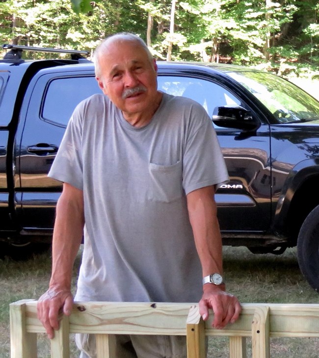 Volunteer Dave Card A man in a gray shirt stands with his hands resting on a wooden bike rack. There is a black truck behind him.