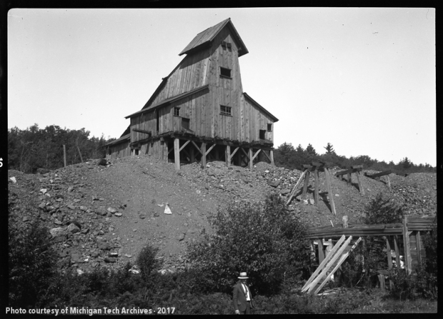 A black and white photo of a wooden industrial building sitting on top of a large pile of rocks. Small shrubs and a man make up the foreground.