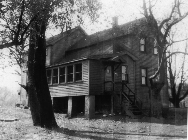 Delgado House at Rockside courtesy Josephine Moss Black and white photo of a large, 2-story, wooden house with a complex roof, surrounded by tall trees. The front door leads into an enclosed porch with a row of 5 windows. The porch is raised up high on cement blocks.