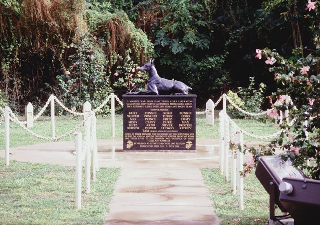 Color photo of a granite memorial with the dogs’ names inscribed on it. A statue of a Doberman sits on top, looking alert. A fence surrounds the memorial. There are trees and flowering bushes in the background.