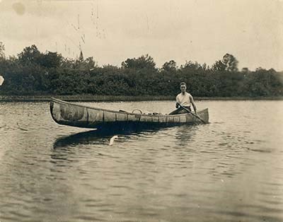 lt= A young man (Franklin Roosevelt) navigating a bay in a birchbark canoe.