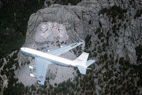 A long white plane with Air Force markings flies over Mount Rushmore, a large carving in the side of a mountain of four faces.
