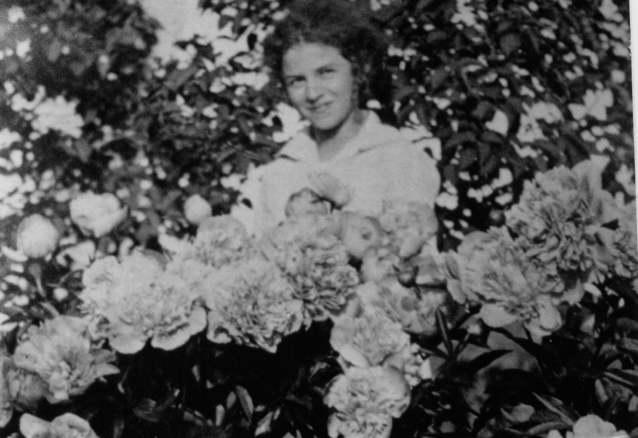 A black and white photo of a woman smiling behind a mound of flowers.
