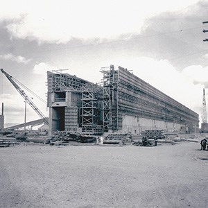 A black and white photo of a large, rectangular structure under construction. The structure is similar to an ocean liner’s hull.