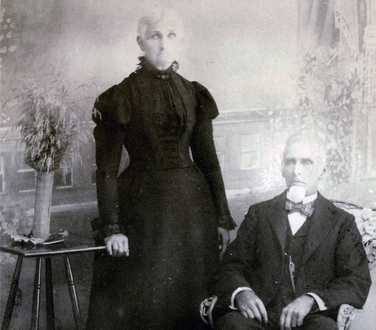 Woman in high-neck black dress stands in portrait next to seated man in suit.