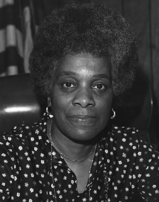 Black and white portrait of Georgia Ellard wearing a dark blouse with a flower pattern. US flag behind her.