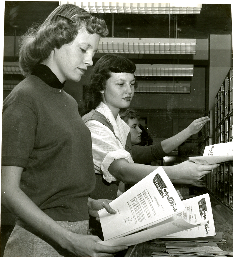 Black and white photo of two white women in sweaters sorting stacks of papers.