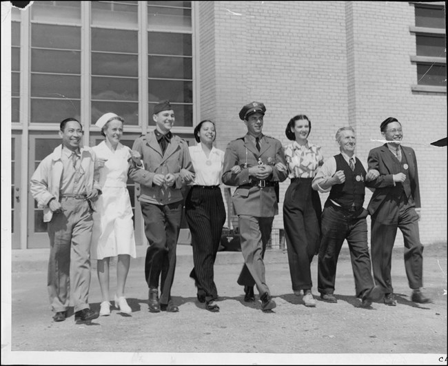 Eight defense workers from different racial, ethnic, gender, and age groups walk arm-in-arm in front of large industrial building.