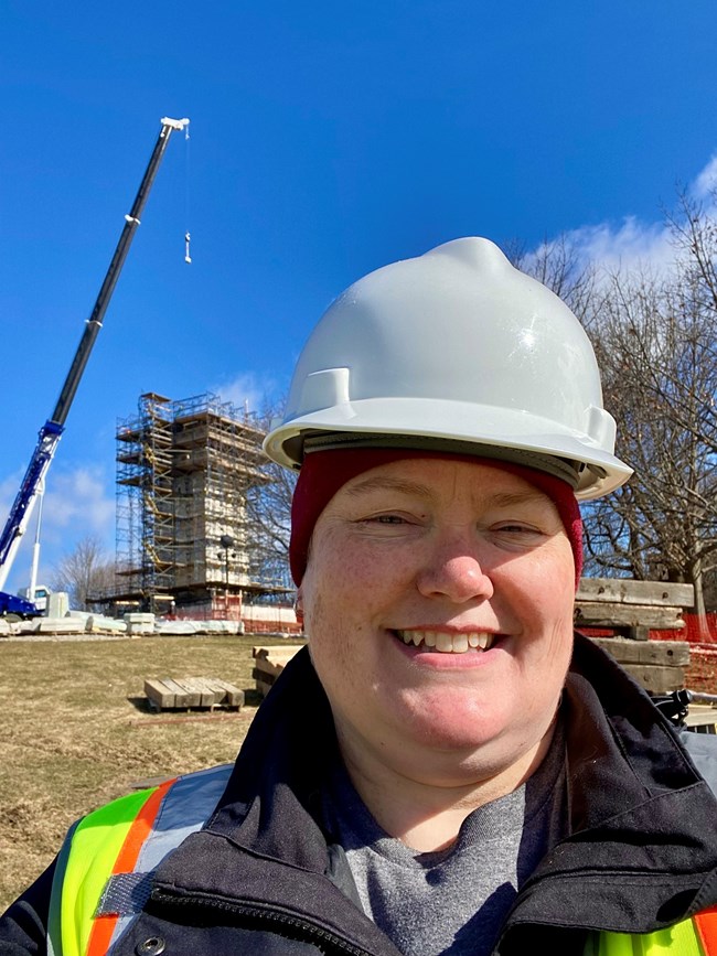Christina smiles for a selfie with a white hardhat on and a building with scaffolding and crane working in the background.
