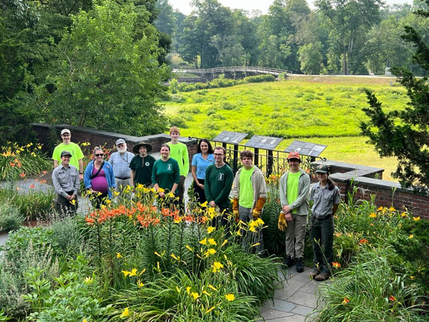 12 individuals stand in a garden with yellow, green, and orange plants. There is a mix of park staff in their gray and green uniforms, young workers in neon green shirts and work pants, some in green shirts, and others in regular street clothes.