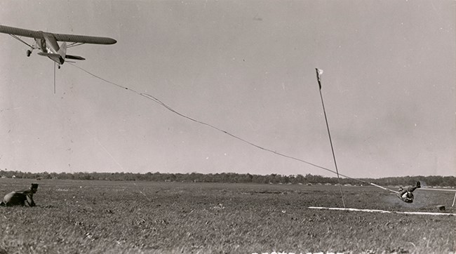 Aircraft takes off from the ground towing a man on a long cable. The man is attached to the cable via a harness and is only a few feet off the ground, having just been picked up.