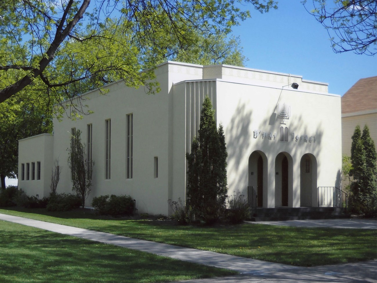 A light-colored building with three arched openings on the front and tall, narrow windows on the side. The words "B'nai Israel" are above the front entrance.
