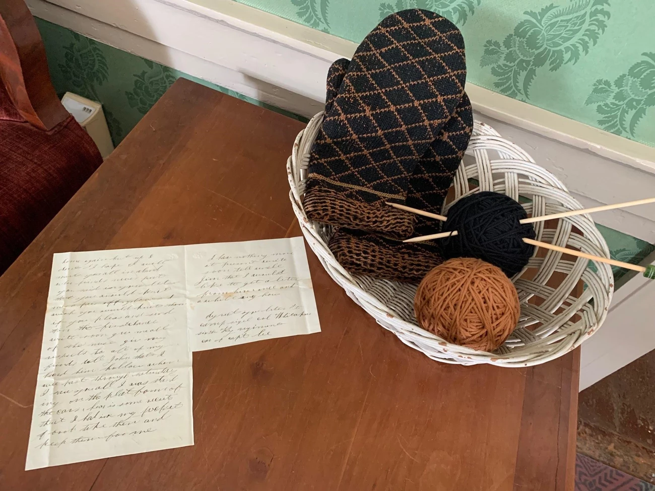 FB Post Mount Parlor 2 Takeover the Network Handwritten letter sits on a table next to a white, wicker basket filled with black and tan yarn, knitting needles, and black and tan knit mittens.