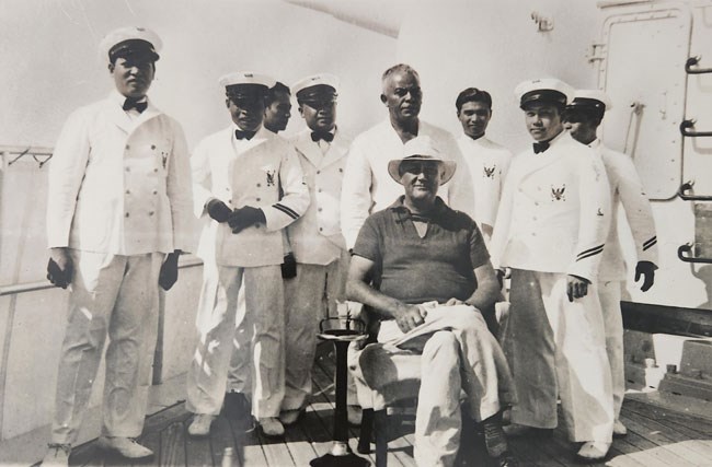 Eight Filipino steward in white uniforms stand around seated FDR aboard a naval ship.