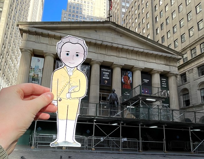 A cartoon illustration of Alexander Hamilton is held up against a backdrop of the balustrades of Federal Hall National Monument.