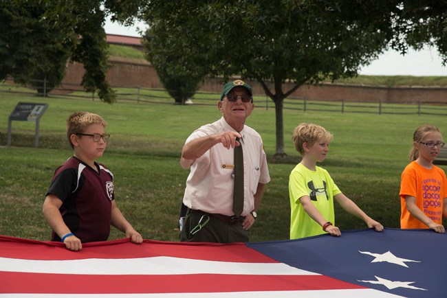 Elderly white man, young white kids on either side of him. They are holding a very large historic replica of the American flag (war of 1812 era). Background is grass with trees.
