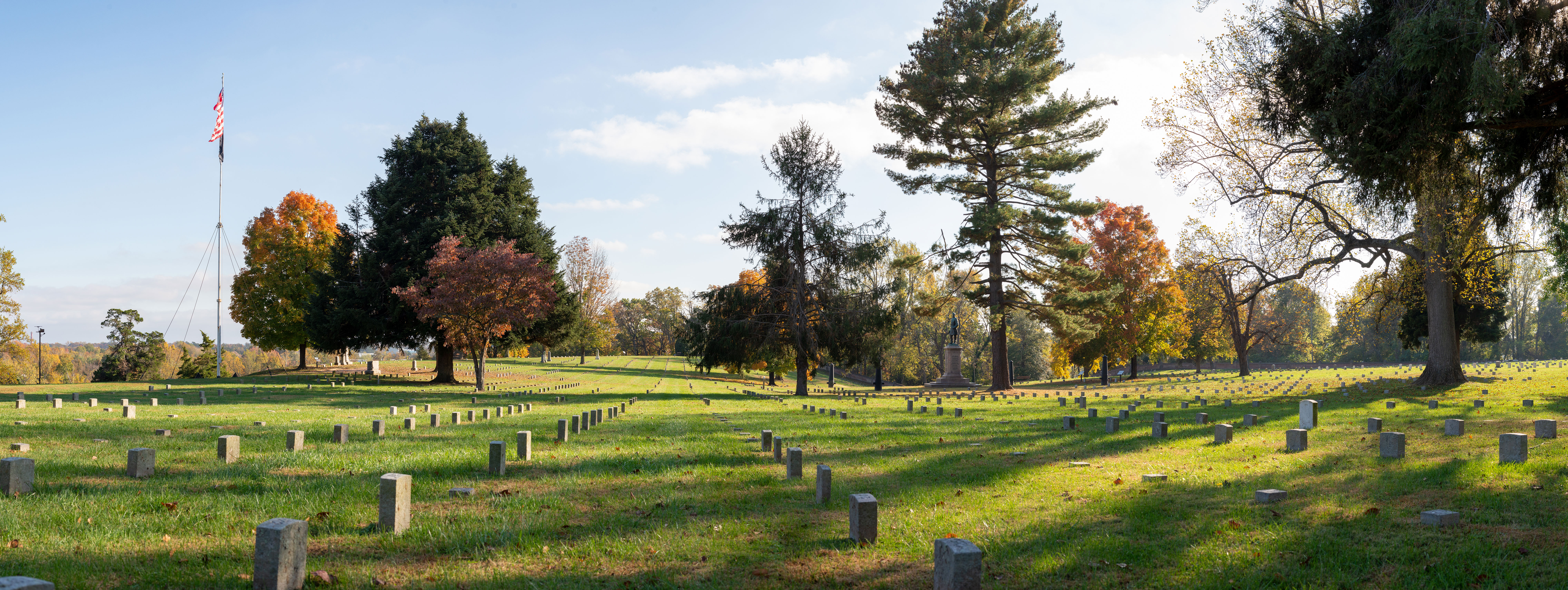 Wide view of national cemetery landscape in fall with flagpole, rows of similar headstones, and scattered trees and monuments