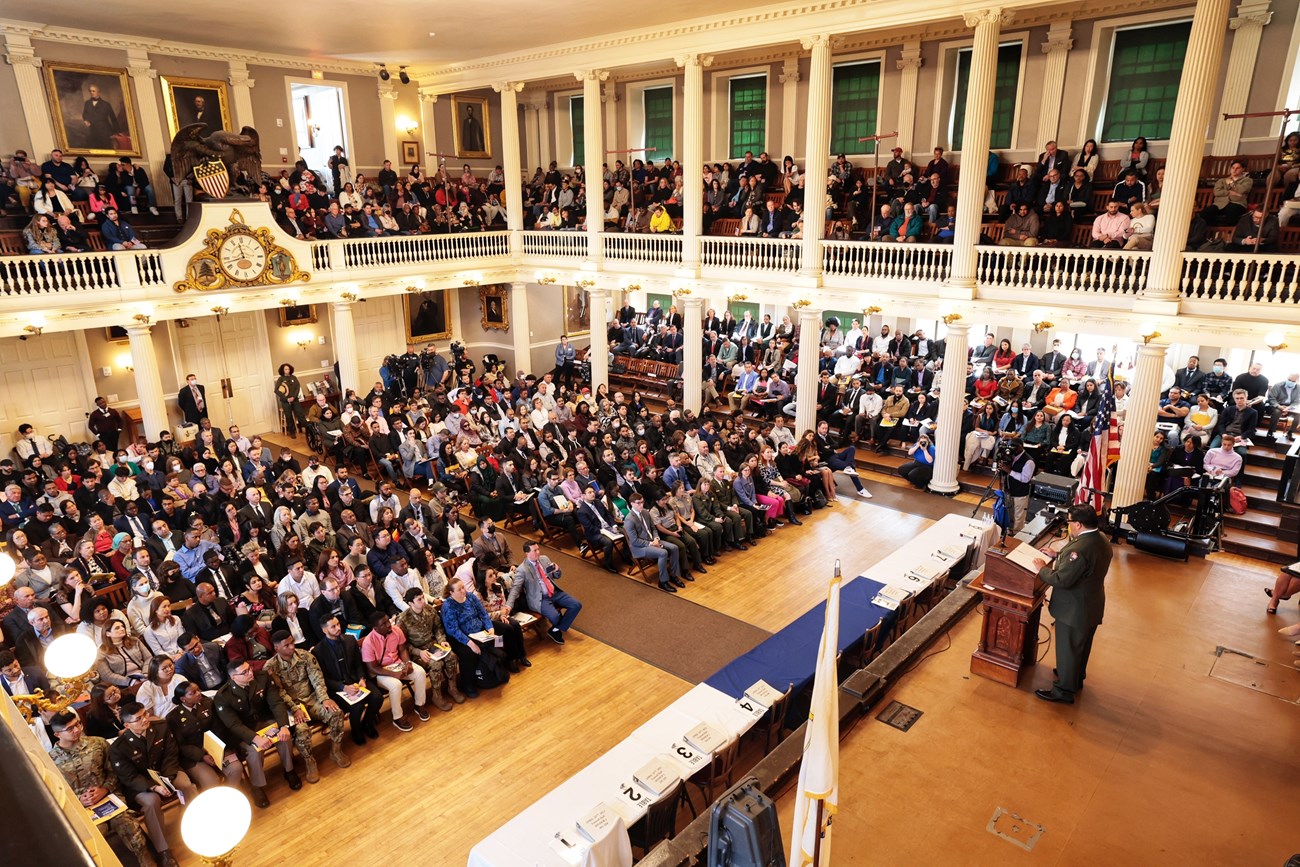 person standing at podium on a stage in front of a meeting hall packed with people.