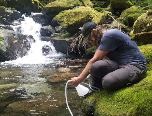 A person sitting on a mossy creek bank collects water with a white flask and plastic tubing.