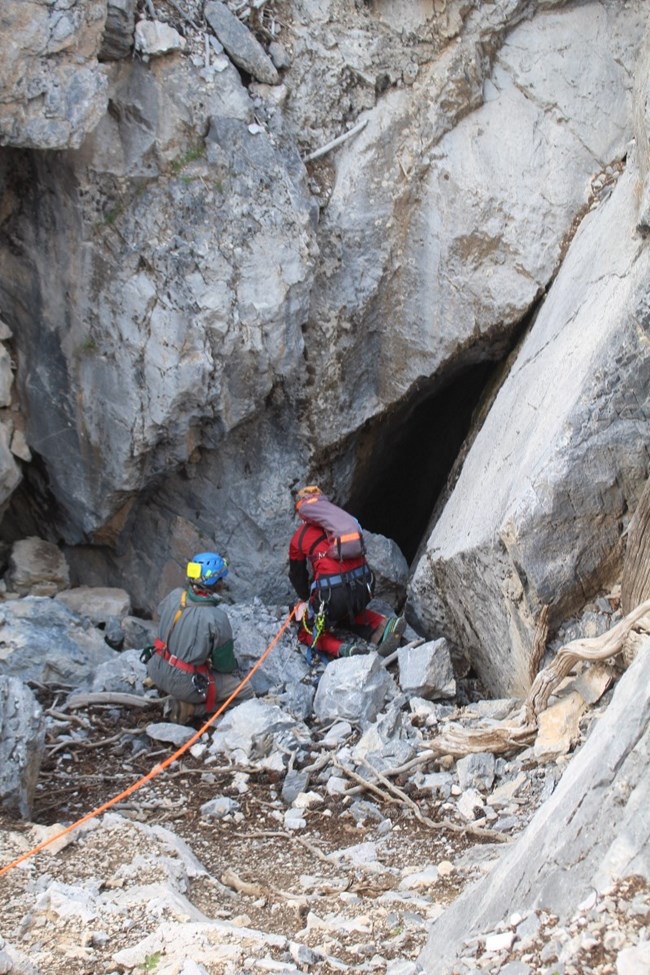Two people and rope at entrance to a hole in ground.