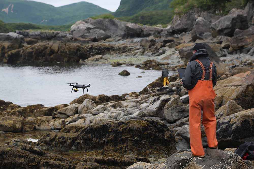 UAS on coast_web A man in orange waders operates a UAS on a rocky coast.