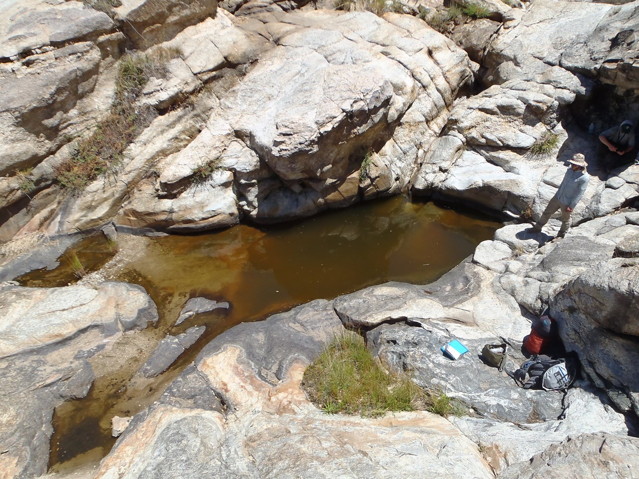 Two people and backpacking and science gear beside a large pool of clear water in a bedrock basin.