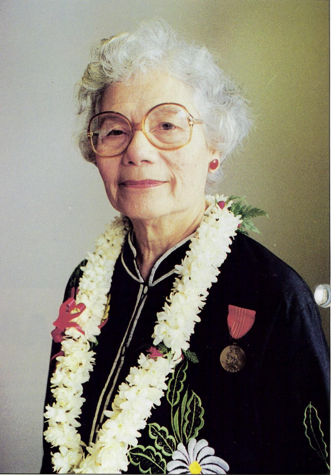 Color photo of an Asian American woman with white hair and glasses. She is wearing a lei and a small medal with a red ribbon.