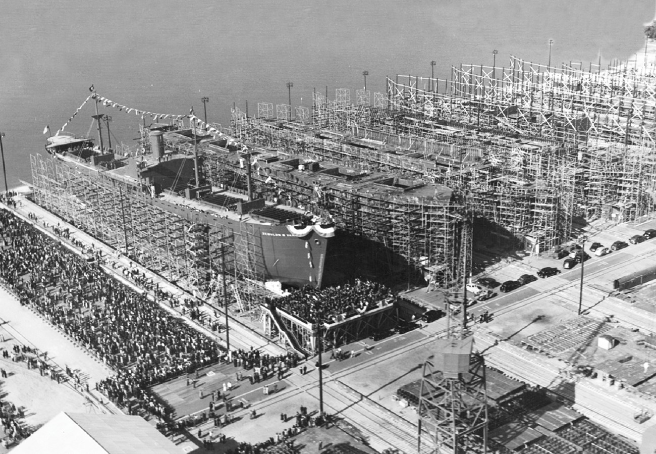 large navy ships lined up at shipyard. crowd is gathered to launch one of the ships.