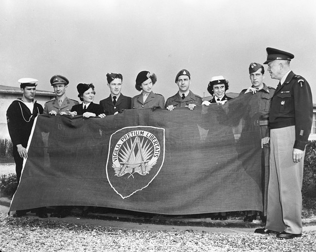 Eisenhower and NATO Flag A black and white image showing General Eisenhower and a row of people in uniforms holding the first NATO flag